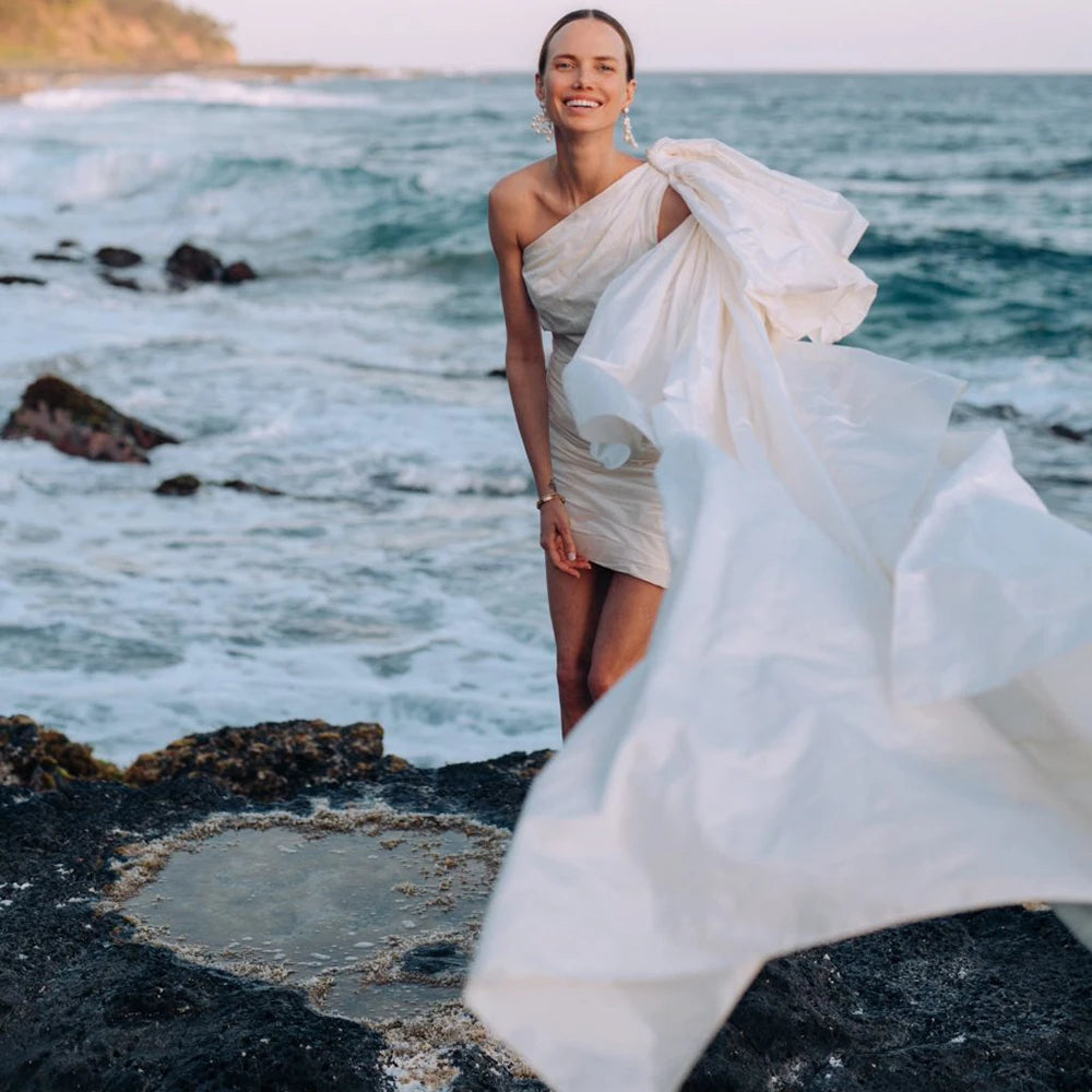 Elevate your wedding look with Robe Elinor. This one-shoulder taffeta dress boasts a chic and elegant design, perfect for a beach wedding. The ruched detailing flatters every curve, while the large bow train adds a touch of sophistication. With its above-knee length, this dress is the epitome of simple yet stylish bridal gowns.